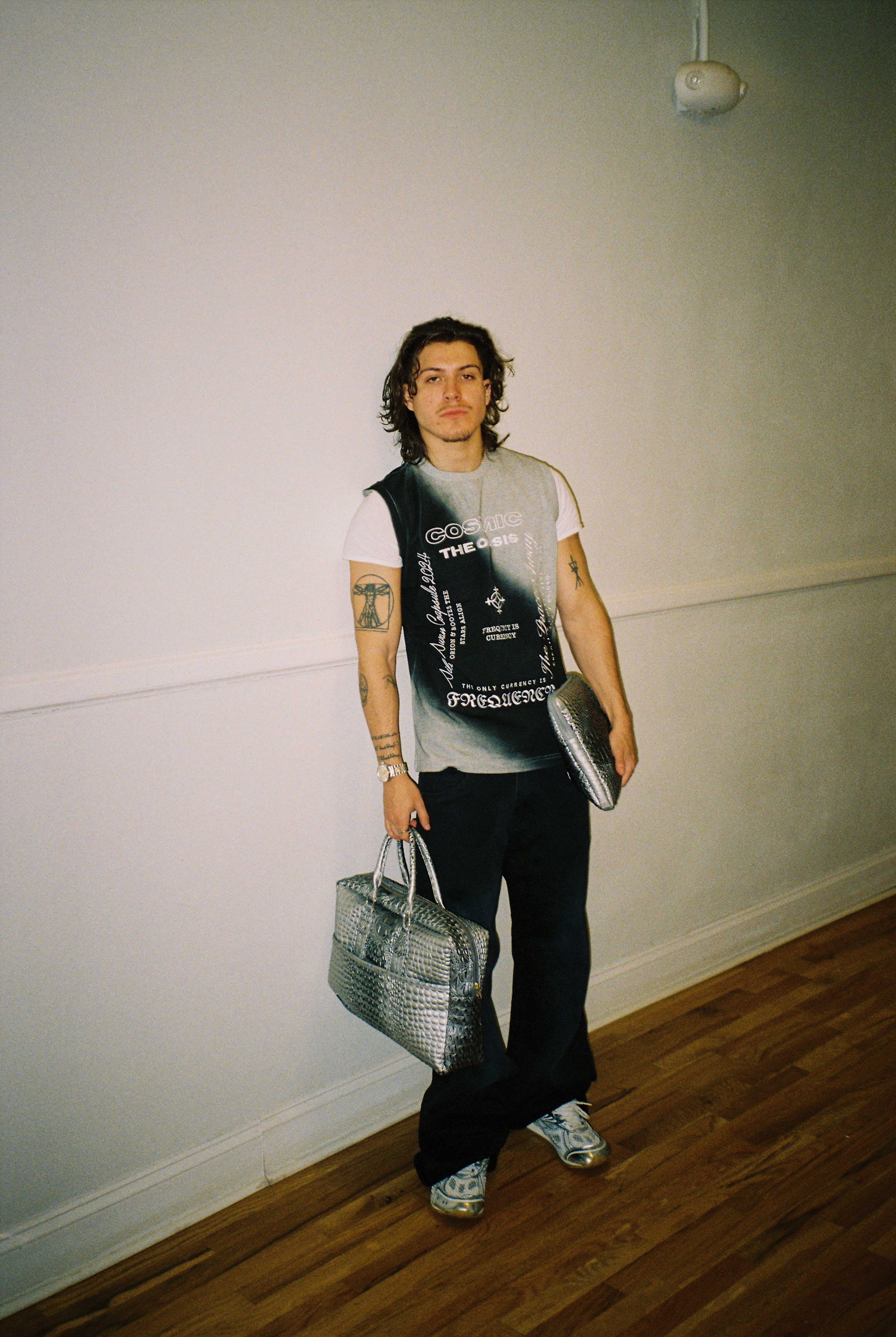 Person holding a silver handbag against a plain wall with a sleeveless shirt.