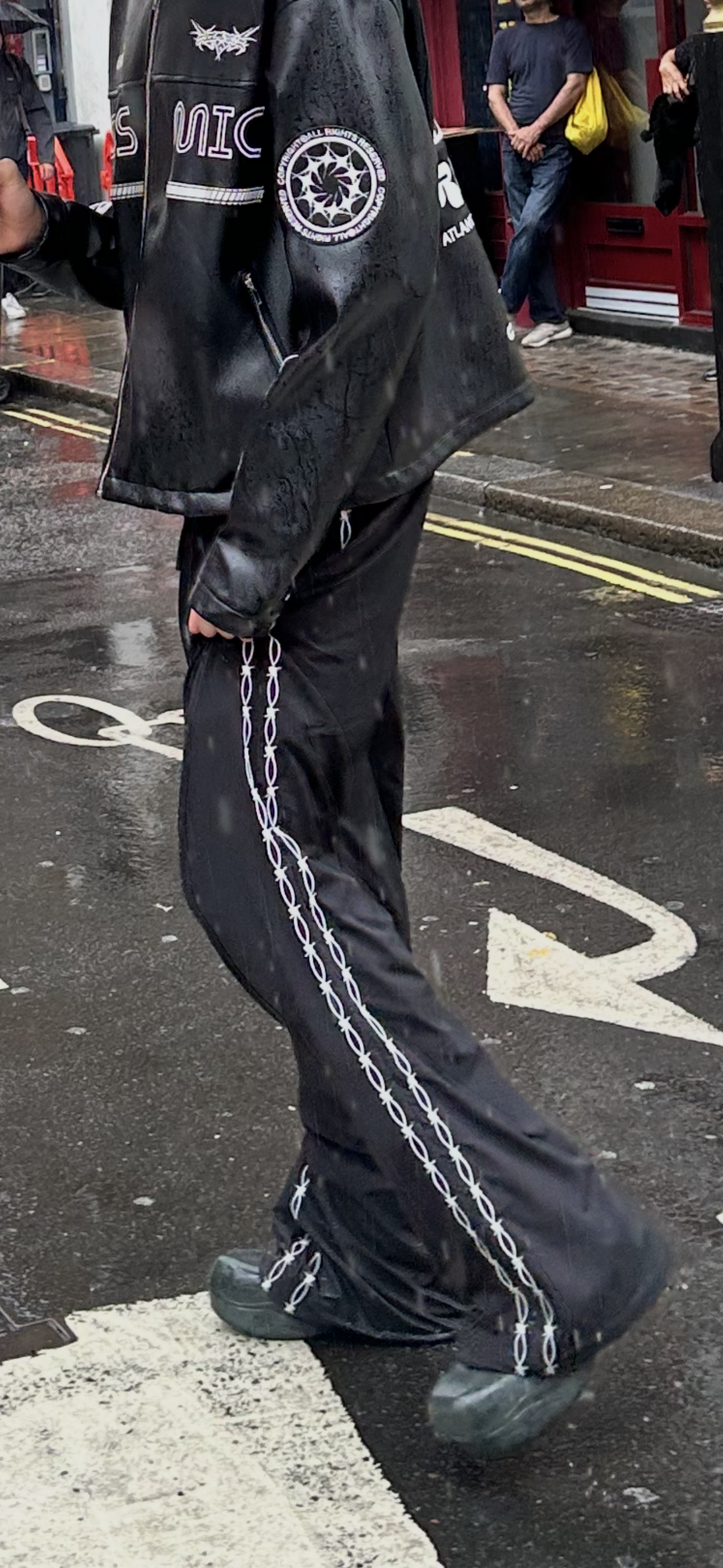 Person wearing a black jacket with logos and black pants with white stripes on a street.